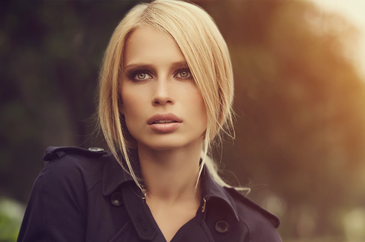 A person with blonde hair and blue eyes wearing a dark coat, looking thoughtfully